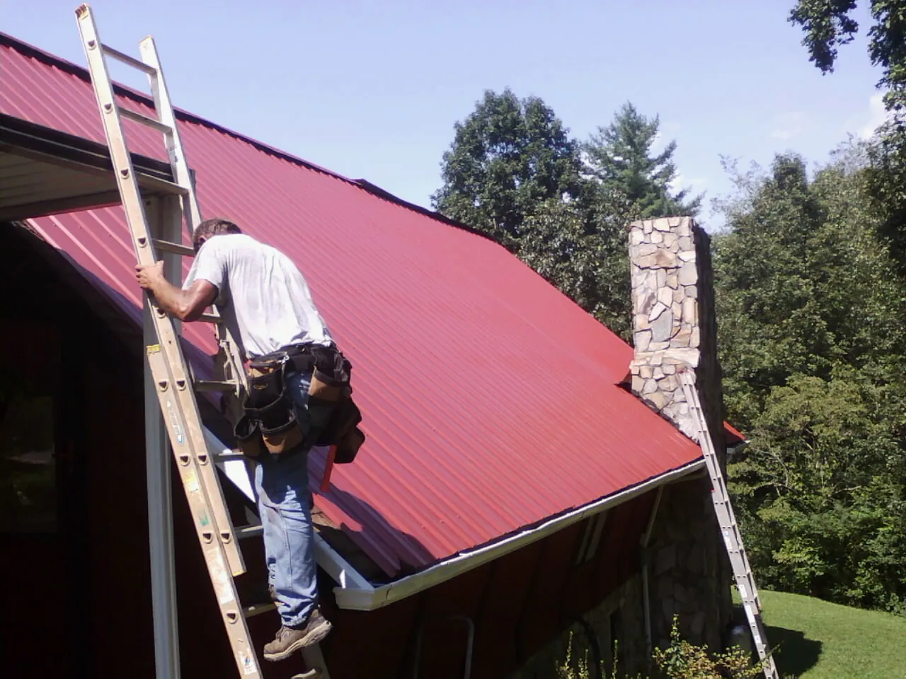Roofer on a ladder beside a red metal roof and stone chimney — Soffit & Fascia Repair by Roofing Brothers in Snoqualmie